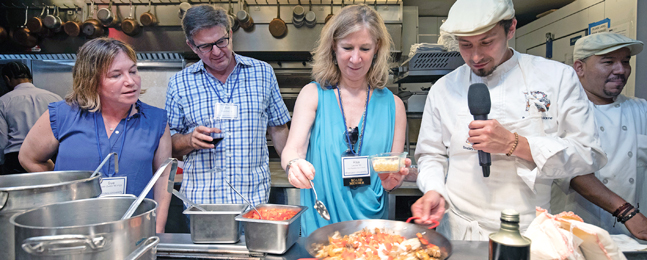 A man in chef's clothes holding a mic with two women and a man stand in a space that looks like an industrial kitchen.